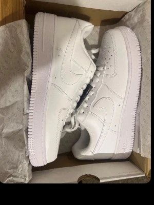 Nike Air Force 1 vita sneakers - Klassiska Nike Air Force 1 sneakers i helvitt läder med vit sula och vita skosnören. Ikonisk design med perforeringar på tån och Nike swoosh på sidan. Perfekta för dig som gillar stilrena och tidlösa sneakers.