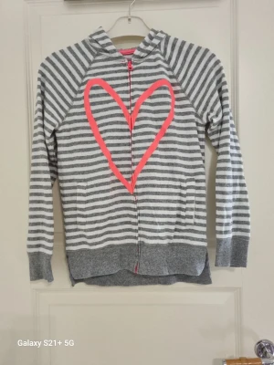 Gråvit randig hoodie med rosa hjärta - Säljer en grå och vit randig hoodie med dragkedja och huva. På framsidan finns ett stort rosa hjärta som ger en cool kontrast. Hoodien har ribbade muddar och är tillverkad i mjukt bomullsmaterial. Perfekt för dig som gillar en avslappnad och trendig stil.