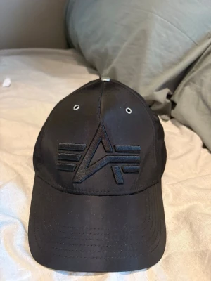 Svart keps med broderad logga - Snygg svart keps med böjd skärm och broderad logga framtill i samma färg. Kepsen har metallknapp på toppen, ventilationshål och justerbar rem bak. Materialet ser ut att vara polyester eller nylon för en sportig känsla.