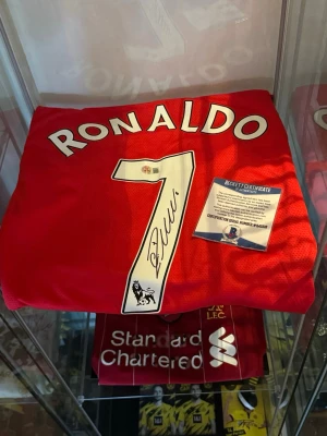 100% äkta Cristiano Ronaldo autograf (signerad Manchester United tröja) med genuint äktenhetsbevis från Beckett - Säljer en 100% äkta Cristiano Ronaldo autograf (signerad Manchester United tröja) med genuint äktenhetsbevis från Beckett. (Beckett är 1 av 3 seriösaste äktenhetsbevis företagen i världen när det kommer till autentisering av autografer) äktenhetsbeviset är ett klistermärke med ett unikt serie nummer och QR kod för ju denna autograf. Signerade Cristiano Ronaldo tröjor är värderade till 15-20 000kr i dagsläget men värdet går upp varje dag! Säljs för endast 8999kr vid en snabb affär.. Mvh Elliot 