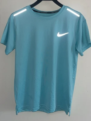 Ljusblå Nike t-shirt med reflexdetaljer - Nike t-shirt i ljusblått med reflekterande detaljer på axlarna och klassisk Nike-logga på bröstet. Tillverkad i ett lätt och ventilerande material, perfekt för träning. Rund halsringning och korta ärmar ger en sportig look.