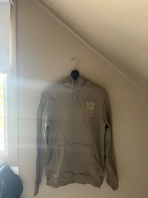 Olivgrön hoodie från Morris - Säljer en olivgrön hoodie från Morris med guldfärgat broderat emblem och liten brittisk flagga på bröstet. Klassisk känguruficka framtill och justerbar huva med snören. Perfekt för dig som gillar stilrena och bekväma plagg.