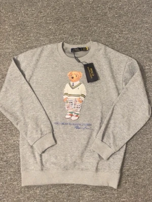 Grå sweatshirt Polo Bear Ralph Lauren - Grå sweatshirt från Ralph Lauren med ikoniskt Polo Bear-tryck framtill. Tröjan har rund halsringning, ribbade muddar och är tillverkad i mjuk bomullsblandning. Perfekt för en avslappnad och trendig look.