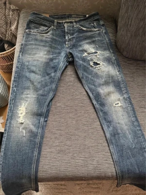 Dondup jeans  - Säljer ett par blå Dondup jeans med slitningar och hål på både fram- och baksida. Jeansen har klassisk femficksmodell och raka ben. Materialet är denim och de har en cool, avslappnad look med tydliga slitdetaljer.