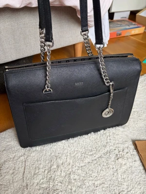 Svart datorväska från DKNY - Stilren svart datorväska från DKNY i strukturerat konstläder. Väskan har dubbla handtag med silverfärgade kedjedetaljer och en dekorativ logotypbricka. Utsidan har en praktisk ficka och insidan är fodrad med DKNY-mönster. Perfekt för laptop och vardagsprylar.