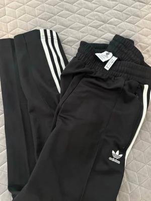 Svarta Adidas mjukisbyxor med vita ränder - Säljer ett par svarta Adidas mjukisbyxor med klassiska vita ränder längs benen och Adidas-logga på ena benet. Byxorna har resår i midjan och dragkedjefickor. Tillverkade i Primeblue-material som innehåller återvunnen plast.