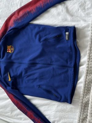 Blå Nike FC Barcelona träningsjacka - Snygg blå träningsjacka från Nike med FC Barcelona-logga på bröstet. Jackan har röda detaljer längs ärmarna och dragkedjeförsedda fickor framtill. Perfekt för dig som gillar fotboll och vill ha en sportig look. 2018 modellen svår att få tag på och en riktigt stilren kofta du kan ha både ute och på träningar