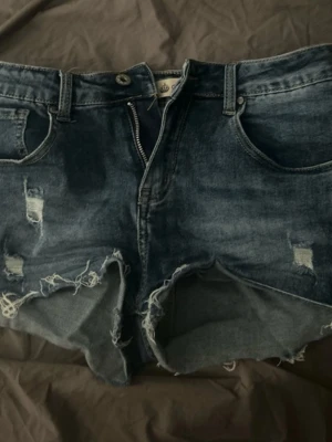 Blå slitna jeansshorts med fransar - Säljer ett par blå jeansshorts med slitningar och fransiga kanter. Shortsen har klassisk femficksmodell, dragkedja och knapp framtill. Perfekta för en avslappnad och trendig look i sommar.