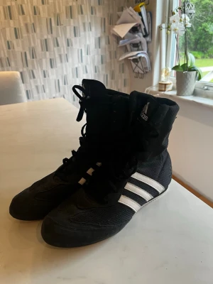 Svarta boxningsskor från Adidas - Säljer ett par svarta boxningsskor från Adidas med klassiska vita ränder på sidorna. Skorna har högt skaft, snörning hela vägen upp och är tillverkade i en kombination av mesh och syntetmaterial för bra ventilation och lätt känsla.