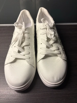 Vita sneakers med klassisk design - Stilrena vita sneakers med lågt skaft och vita snören. Skorna har en enkel och clean look med dekorativa ränder på sidan. Tillverkade i ett slätt syntetmaterial som ger en fräsch känsla. Perfekta för dig som gillar minimalistisk stil.