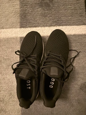 Svarta Adidas Ultraboost sneakers - Svarta vita Adidas Ultraboost sneakers med stickad ovandel och klassiska tre ränder på sidan. Skorna har vita Boost-sulor och svarta detaljer. Perfekta för en sportig och stilren look. Skön passform med snörning och ventilerande material.
