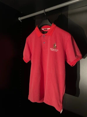 Röd pikétröja från Polo Club Sällsynt - Snygg röd pikétröja från Royal County of Berkshire Polo Club med klassisk krage, knappar och broderad logga på bröstet. Perfekt till jeans eller shorts för en sportig och stilren look.
