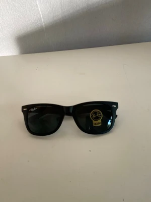 Svarta Ray-Ban Wayfarer solglasögon - Klassiska svarta Ray-Ban Wayfarer solglasögon med bred båge och mörka glas. Ikonisk design med Ray-Ban-logga på linsen och sidan. Passar perfekt för dig som vill ha en tidlös och cool look i solen.