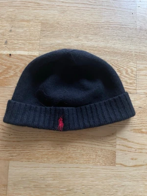 Svart mössa med röd logga - En svart, ribbstickad mössa med uppvikt kant och en liten röd broderad logga framtill. Klassisk och enkel design som passar till det mesta. Mössan är mjuk och ser ut att vara gjord i ull eller ullblandning.