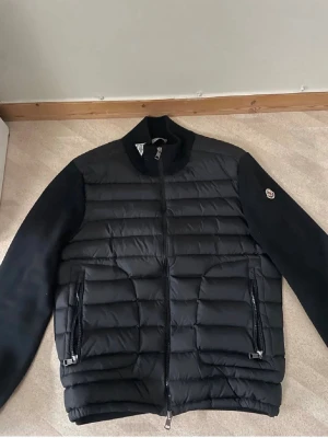 Svart moncler cardigan  - Snygg svart moncler cardigan, STRL L- passar mer till M. med quiltad kropp och släta ärmar. Jackan har två dragkedjeförsedda fickor framtill och hög krage. Moncler-logga på ena ärmen