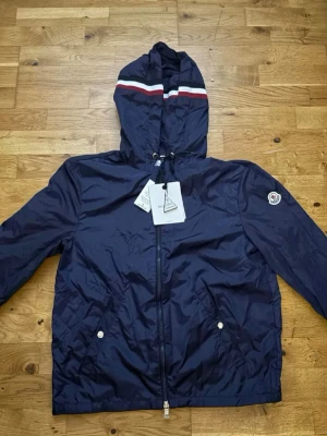 Mörkblå vindjacka från Moncler - Snygg mörkblå vindjacka från Moncler med huva och dragkedja. Jackan har vita och röda ränder på huvan, två fickor med tryckknappar och Moncler-logga på ärmen. Materialet är lätt och vattenavvisande, perfekt för blåsiga dagar.