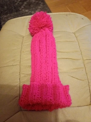 Stickad rosa mössa med tofs stl 4-6 one size - Säljer en lång, ribbstickad mössa i starkt rosa färg. Mössan har en stor bolltofs upptill och uppvikt kant. Perfekt för dig som vill sticka ut lite extra under kalla dagar. Materialet känns mjukt och värmande, troligen akryl eller ullblandning. Hemstickad.