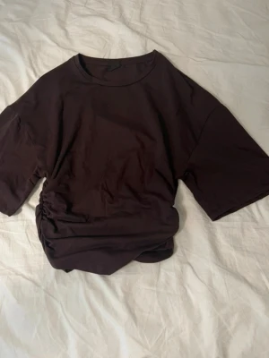 Brun t-shirt med snörning  - Brun t-shirt med korta ärmar och rund hals. Snygg snörningsdetalj i sidan. Tillverkad i mjuk bomull.