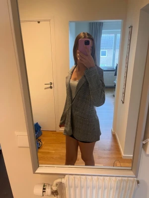 Grå rutig oversized kavaj - Snygg grå kavaj med rutigt mönster och oversized passform. Klassisk krage och två knappar framtill. Perfekt att styla med både jeans och kjol för en trendig look. Materialet ser ut att vara en blandning av polyester och viskos.