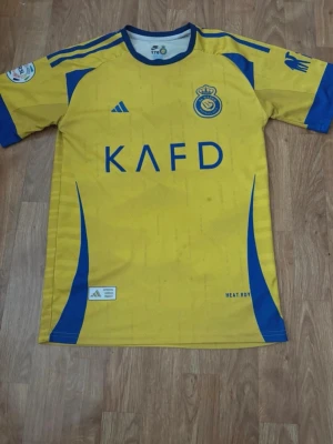 Gul Al Nassr matchtröja Ronaldo 7 - Snygg gul matchtröja från Al Nassr med blå detaljer och tryck. Framsidan har klubbmärke och sponsorlogga, baksidan har namnet Ronaldo och nummer 7. Tillverkad i lätt och ventilerande material, perfekt för fotbollsträning eller match.