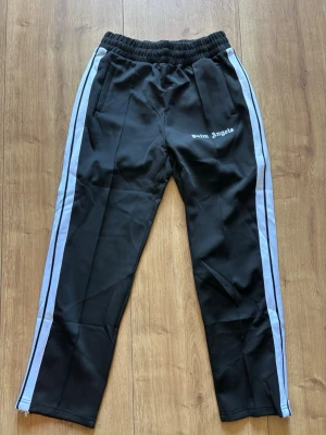 Palm Angels svarta mjukisbyxor - Palm Angels svarta mjukisbyxor med vita sidoränder och logga på vänster sida. Byxorna har resår i midjan och raka ben. Tillverkade i ett glansigt material som ger en sportig vibe. Perfekta för en avslappnad och trendig streetwear-look.