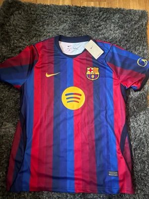 FC Barcelona 26-27 Hemma Tröja - Säljer en FC Barcelona replika matchtröja med klassiska blå och röda vertikala ränder. Tröjan har klubbmärke, Nike-logga och Spotify som sponsor framtill. Tillverkad i lätt och ventilerande material, perfekt för fotboll eller träning. Tröjan står att den är i XL men passar mer L.