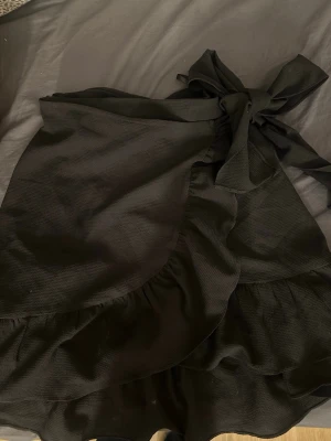 Svart omlottkjol med volang - Säljer en svart omlottkjol med volangkant och knytband i midjan. Kjolen har en lätt struktur i tyget och ger en snygg och trendig look. Perfekt att matcha med en enkel topp eller stickad tröja.