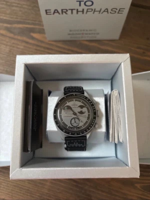 Omega X Swatch  - Stilren klocka i svart och silver med månfasdetalj på urtavlan. Klockan har svart läderarmband med vita sömmar och en tydlig boett. Baksidan är graverad med en måne och texten 'Moon to the moon'. Perfekt för dig som gillar unika detaljer.