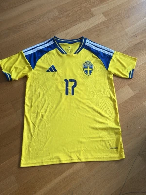 Svensk landslagströja Adidas nr 17 - Gul och blå svensk landslagströja från Adidas med nummer 17 och namnet Gyökeres på ryggen. Tröjan har blå detaljer på axlar och ärmslut, samt broderad landslagssköld på bröstet. Tillverkad i lätt och ventilerande material, perfekt för fotboll.