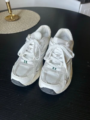 Vita sneakers med gröna detaljer från H&M stl 37 - Säljer ett par vita sneakers från H&M i stl 27  med mesh och skinnpaneler samt gröna detaljer på sidan och tungan. Skorna har klassisk snörning och en chunky sula som ger en sportig look. Perfekta för dig som gillar retrostil och bekväma skor.