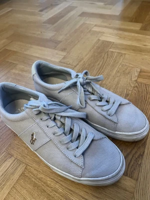 Ljusgrå sneakers från Polo Ralph Lauren - Snygga ljusgrå sneakers från Polo Ralph Lauren med klassisk låg profil och canvasmaterial. Skorna har vita sulor, matchande ljusgrå skosnören och den ikoniska Polo-loggan i guld på sidan. Perfekta för en clean och stilren look. Säljs då de inte används