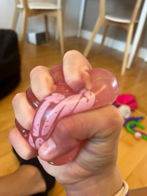 Rosa squishy stressboll - En rosa squishy stressboll fylld med glitter och små vita partiklar. Bollen är mjuk och formbar, perfekt att klämma på när man vill slappna av eller behöver något att pilla med. Tillverkad i elastiskt gummiliknande material.