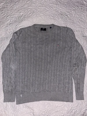 Gant  - Snygg grå kabelstickad tröja med rund hals och ribbade muddar. Tröjan har ett diskret broderat märke på bröstet och är perfekt för lager på lager. Passar till både jeans och kjol för en chill look.