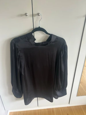 Svart satinblus med puffärm - Stilren svart blus i glansigt satinmaterial med puffiga långärmar och hög krage. Blusen har en rak passform och breda manschetter som ger en trendig look. Perfekt att matcha med jeans eller kjol för en snygg outfit. Blusen har storlek 50 men är liten i storlek så skulle säga att den är runt 42-44 istället❤️