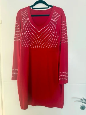 Röd klänning med dekorativa detaljer - ❤️✨ Röd klänning med dekorativa detaljer ✨❤️  Elegant röd klänning med långa ärmar och vackra dekorativa stenar som bildar geometriska mönster ✨ En stilfull modell som passar både till vardag och fest ❤️  🌸 Skick: mycket fint — endast använd en gång 🤍  📏 Mått: • bredd: 116 cm (kan töjas till ca 125 cm) • längd: 91 cm  📌 Storlek: L/XL