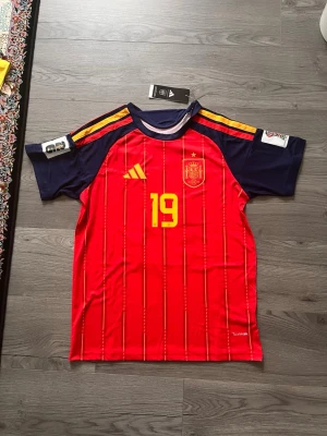 Spaniens matchtröja Adidas röd - Säljer en officiell röd matchtröja från Spaniens landslag med gula detaljer och nummer 19 på bröstet. Tröjan har mörkblå ärmar, gula ränder på axlarna och landslagets emblem på bröstet. Perfekt för fotbollsfans och samlare.