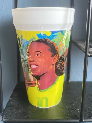 Ronaldinho McDonalds kopp - Färgglad plastmugg med illustration av en fotbollsspelare i gul tröja och nummer 10. Muggen har livliga färger som blått, gult och grönt och är dekorerad med sporttema. Perfekt för drycker eller som samlarobjekt.