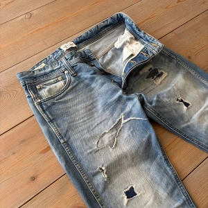 Vintage jeans från Jack n’ Jones -  snygg look👖  - Blå anti fit jeans från Jack n’ Jones i strl 29/32. Sitter som Slim fit och har en sliten look - står ut i mängden. Har igen sydda hål och några fläckar men jeansen är fortfarande väldigt snygga. 🪡Saknar en knapp (se bild) - går att laga🎞️Klassiska vintagejeans 🙌Snygg passform Endast 200!