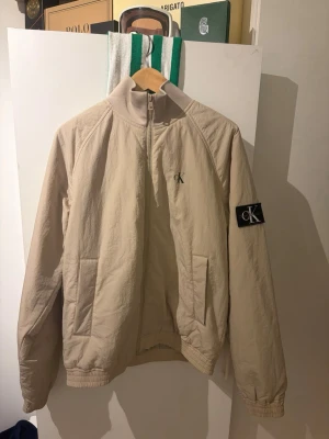 Beige vindjacka från Calvin Klein - Stilren beige vindjacka från Calvin Klein med dragkedja, hög krage och två fickor framtill. Diskret CK-logga på bröstet och en större patch på ärmen. Jackan har resår vid ärmslut och midja för en snygg passform. Perfekt för blåsiga dagar. Jackan är aldrig använd. ABS! Pris går absolut att diskutera!