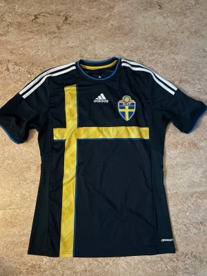 Svensk landslagströja Adidas blå - Säljer en svart svensk landslagströja från Adidas med gult kors och landslagsemblem på bröstet. Tröjan har korta ärmar, vita ränder på axlarna och är tillverkad i polyester. Saknar storleks anvisningar men passar xs/s