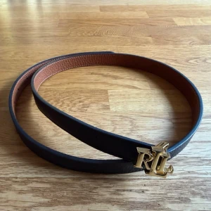 Vändbart skärp med Lauren Ralph Lauren-spänne - Snyggt vändbart bälte i skinn med ena sidan svart och andra sidan brun. Spännet är i guld med Lauren Ralph Lauren-logga framtill. Ordinarie pris 1200kr.