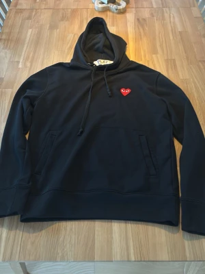 Svart hoodie från CDG - Svart hoodie med huva och dragsnören, samt en ficka framtill. På bröstet finns ett litet rött hjärta med ögon som detalj. Hoodien har en klassisk passform och är tillverkad i mjukt material, perfekt för en avslappnad stil.
