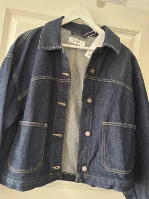 Mörkblå oversized jeansjacka - Snygg mörkblå jeansjacka i oversized modell med klassisk krage och stora framfickor. Jackan har synliga kontrastsömmar och knäppning framtill med metallknappar. Perfekt för en avslappnad och trendig look.