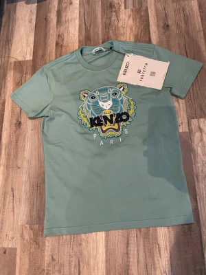 Grön Kenzo t-shirt med tigerbrodyr - Snygg grön t-shirt från Kenzo med ikonisk tigerbrodyr och texten 'Kenzo Paris' på bröstet. T-shirten har rund hals och korta ärmar. Perfekt för dig som vill sticka ut med en trendig och unik look.Kommer med autenticitet kort SKICKAS INNAN TISDAG DEN 21 Sitter tajt runt armar o bröst o löst runt magen