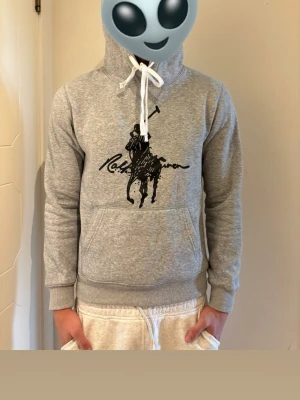 Grå hoodie Ralph lauren - Snygg grå hoodie med stor svart Polo-inspirerad logga på bröstet och vit dragsko i huvan. Klassisk känguruficka framtill och ribbade muddar vid ärmslut och midja. Perfekt för en avslappnad och trendig look.