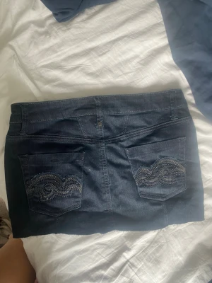 Mörkblå jeanskjol med broderade fickor - Snygg mörkblå jeanskjol i kort modell med klassiska fickor fram och bak. Bakfickorna har dekorativa broderier som ger kjolen en unik touch. Perfekt för dig som gillar denim och vill ha en enkel men cool stil.