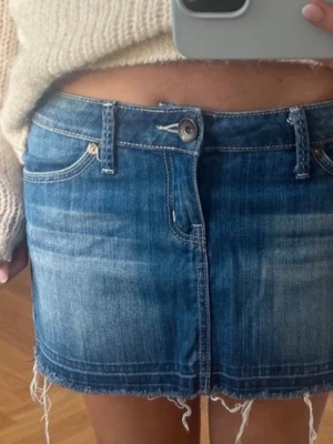 Blå jeanskjol  - Säljer en blå kort jeanskjol med rå, fransig kant och slitna detaljer. Kjolen har klassiska fem fickor, bälteshällor och knappstängning framtill. På ena bakfickan finns en broderad detalj i guld. 