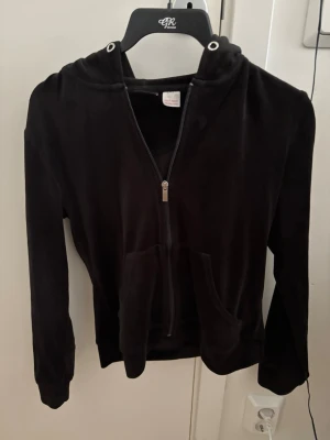Svart velour hoodie med dragkedja - Mjuk och skön svart hoodie i velour med dragkedja framtill och två fickor. Tröjan har huva med snörning och långa ärmar. Perfekt för en avslappnad och trendig look.