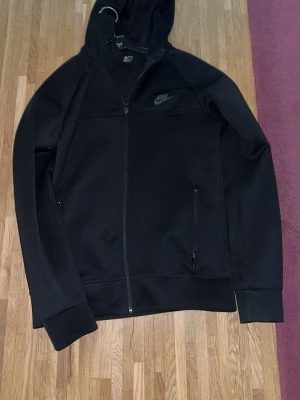 Svart Nike hoodie med dragkedja - Säljer en svart hoodie från Nike med hel dragkedja framtill och två fickor med dragkedja. Klassisk Nike-logga på bröstet och huva med snörning. Tillverkad i mjukt material som passar perfekt till en avslappnad stil.