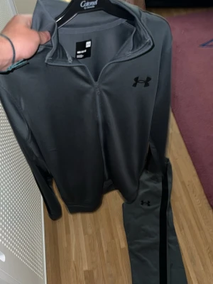 Mörkgrå träningsset från Under Armour - Säljer ett mörkgrått träningsset från Under Armour i storlek M. Setet består av en zip-jacka med hög krage och matchande byxor med svarta detaljer längs sidorna. Materialet är mjukt och stretchigt, perfekt för träning eller chill. Diskret logga på både jacka och byxa.